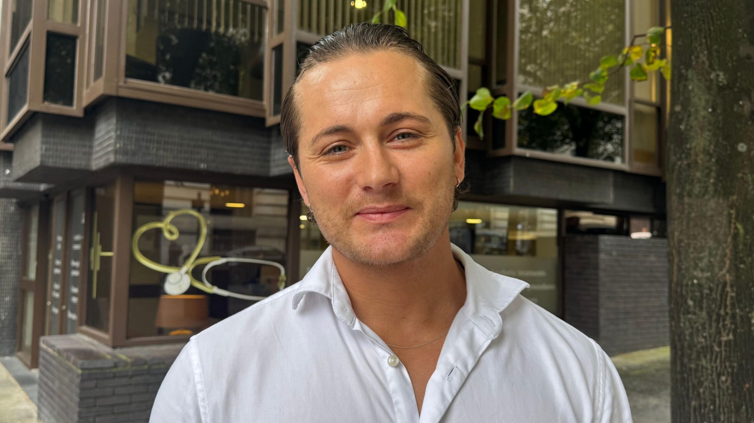 Max Longman, a man in his thirties, with his hair slicked back, wearing a white shirt opened at the collar, standing in front of a building