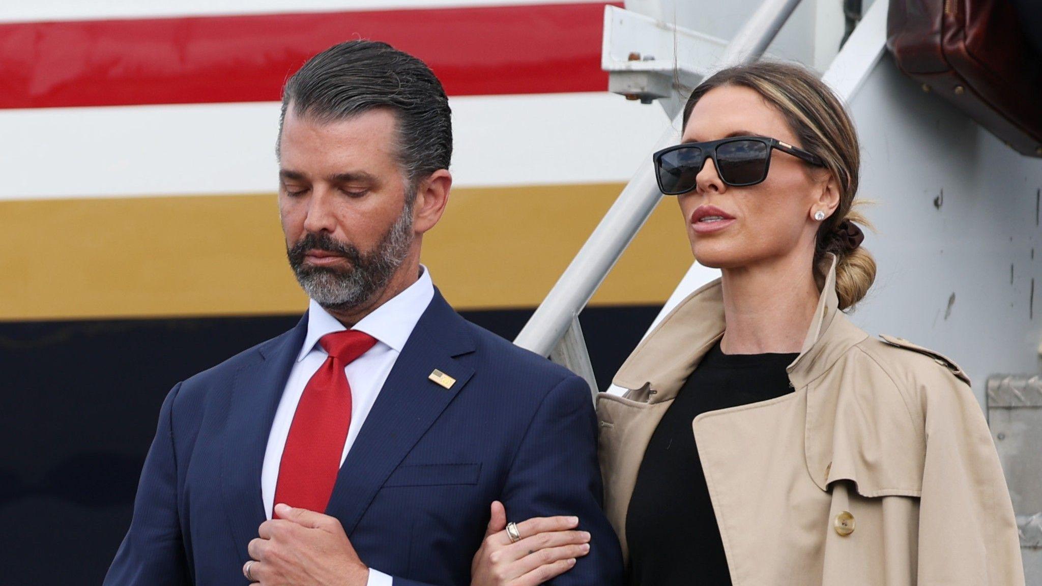Donald Trump Jr. and his wife Bettina Anderson are seen close up with a plane behind them. She is on the right in a blacktop, tan trench coat and black sunglasses. Her hair is pulled back. He is wearing a blue suit and red tie.