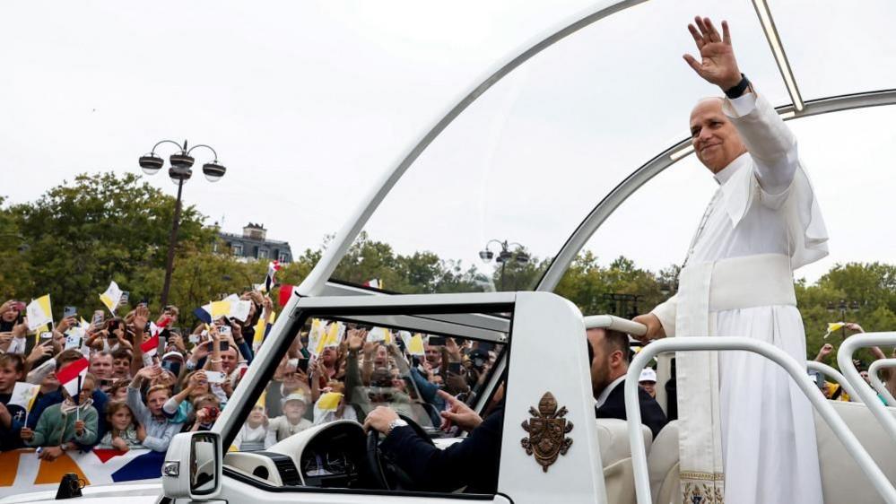 Pope Leo smiles and holds his arm up in his popemobile. Crowds can be seen waving flags and gesturing towards the pope. Some are holding up their phones or cameras to take pictures
