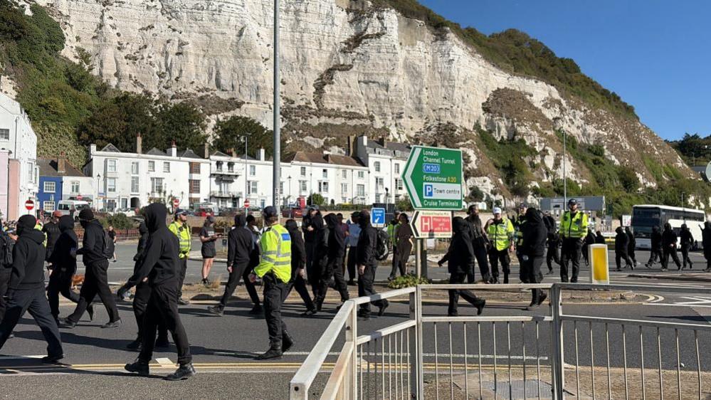 A group of people in black clothing wearing black balaclavas. A number of police officers are in the crowd.