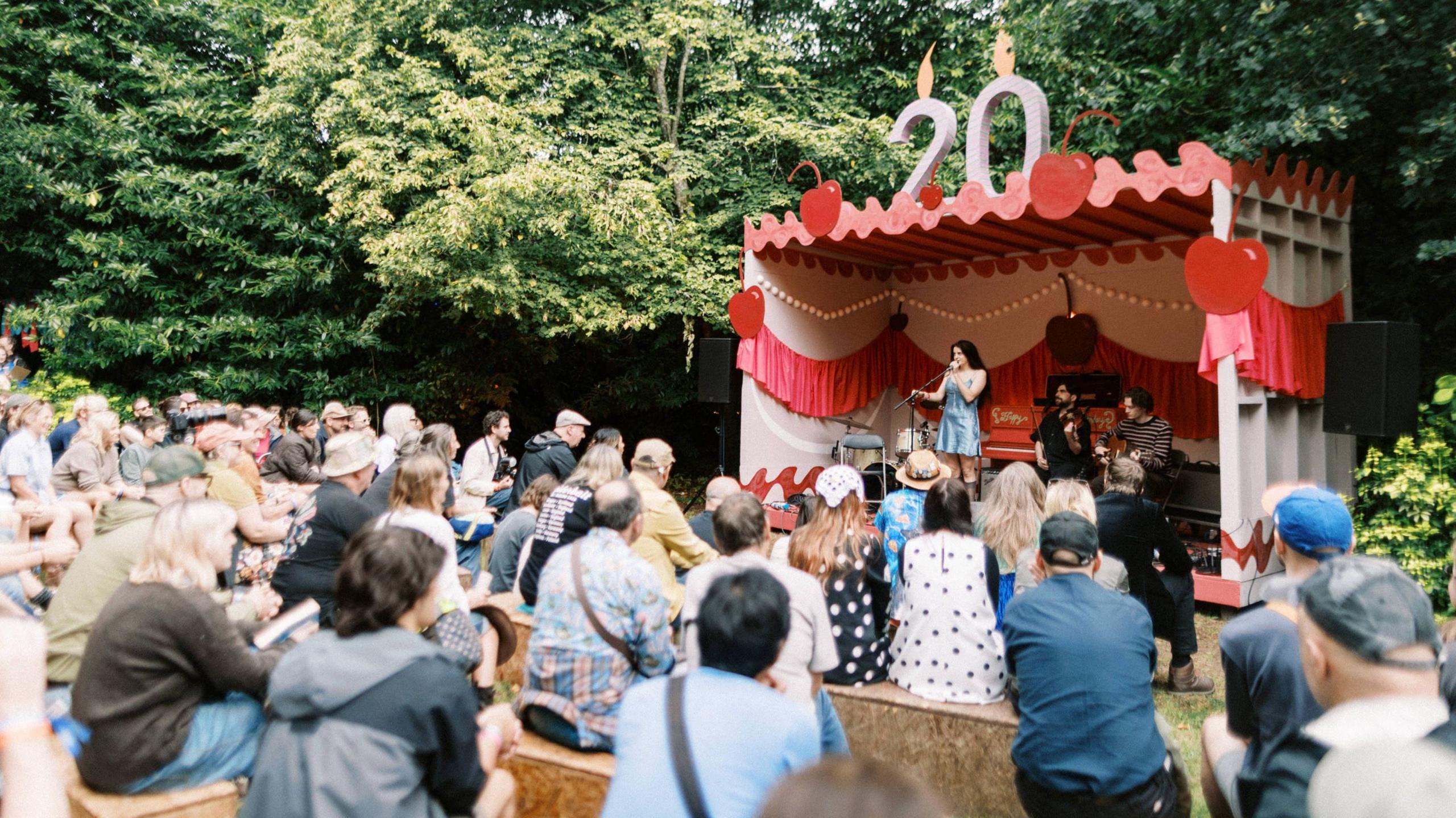 A crowd of people watching a woman on stage in a daytime performance in a wooded area.