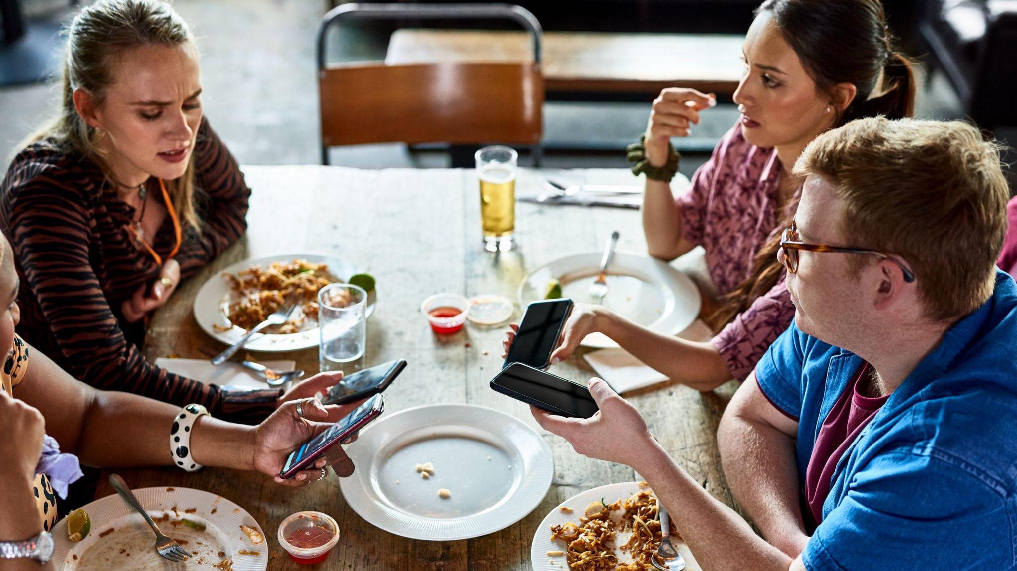 Three young people viewed from above are sitting at a table at the end of a meal. They have their phones out and the woman on the left is frowning as she looks at hers. There are empty and half empty plates and glasses.