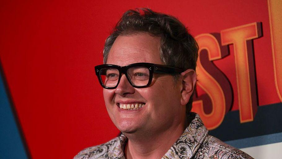 Comedian Alan Carr with his trademark large glasses in a colourful shirt against a red background