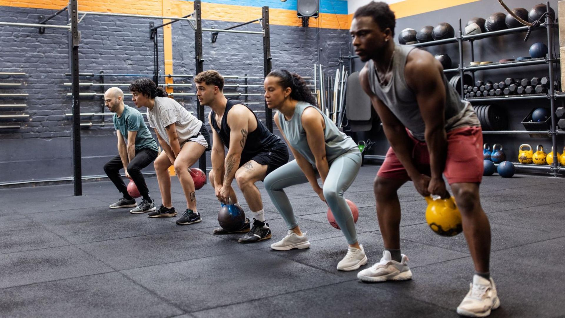 People in a gym are lined up using kettle bells, swinging them between their legs with feet apart - behind them are shelves of weights including dumb bells and bar bells. The walls are painted grey. The floor mats are also grey.