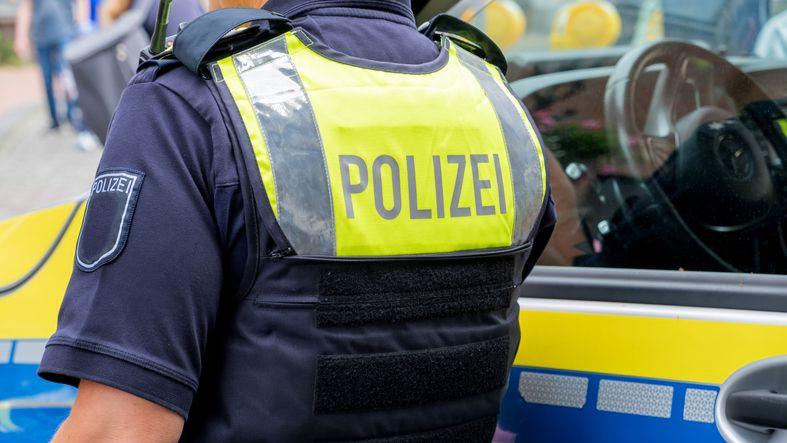 A German police officer standing next to a police vehicle. The officer wears an operational vest that reads "polizei".