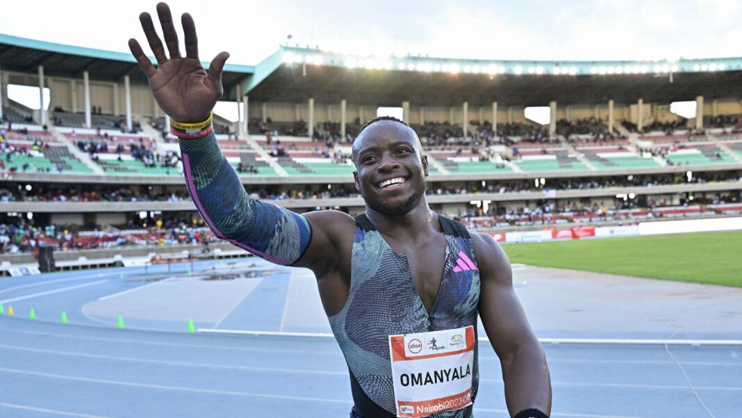 African greats inspired by the potential of Nairobi 2029 Ferdinand Omanyala, visible from waist up wearing matching athletics vest, unzipped over the chest, and sleeve over his right arm, smiles and waves to the crowd with his right arm raised while standing on the blue running track at the Kasarani Stadium. Sparsely populated stands can be seen running around the bend of the track behind him, with floodlights twinkling despite the fact that the sky is not yet dark