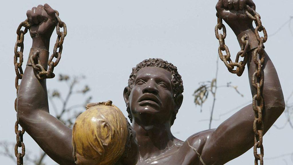 Close up of statue showing liberation from slavery on Goree Island in Senegal. It is a man from shoulders up, with his arms raised, holding a broken chain in his hands which is still attached to his wrists. The statue also includes the figure of a woman standing in front of the man - all that is visible of her is the back of her head, and she has a head wrap on.