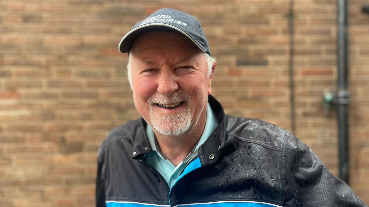 A photo fo a man standing smiling in the camera. He has got a cap on and a rain coat which is black and a blue top on underneath.