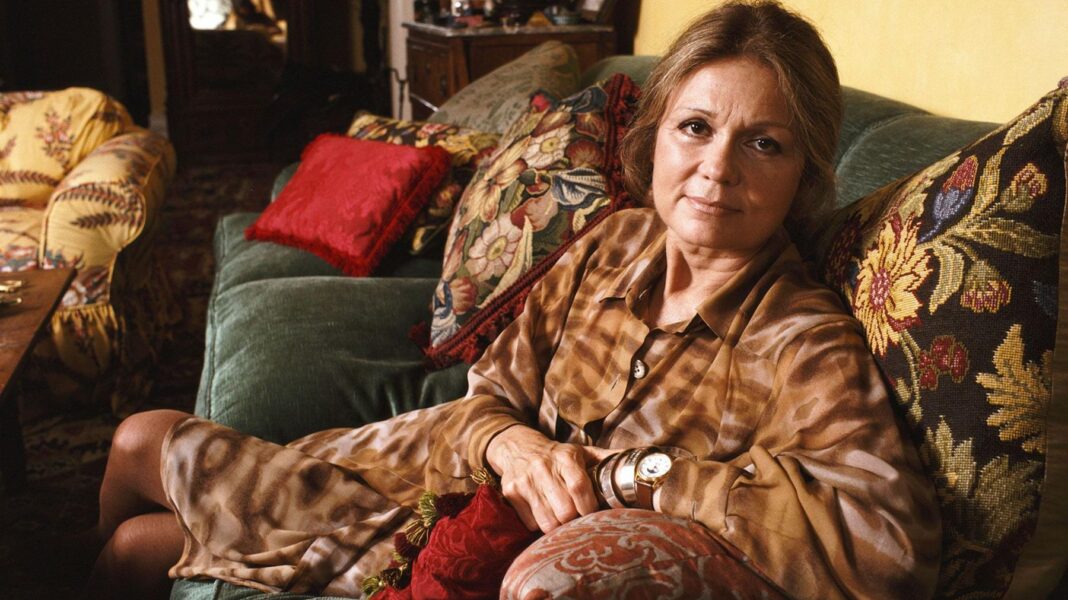 Gloria Steinem lying on a couch in a brown shirt dress surrounded by colourful cushions