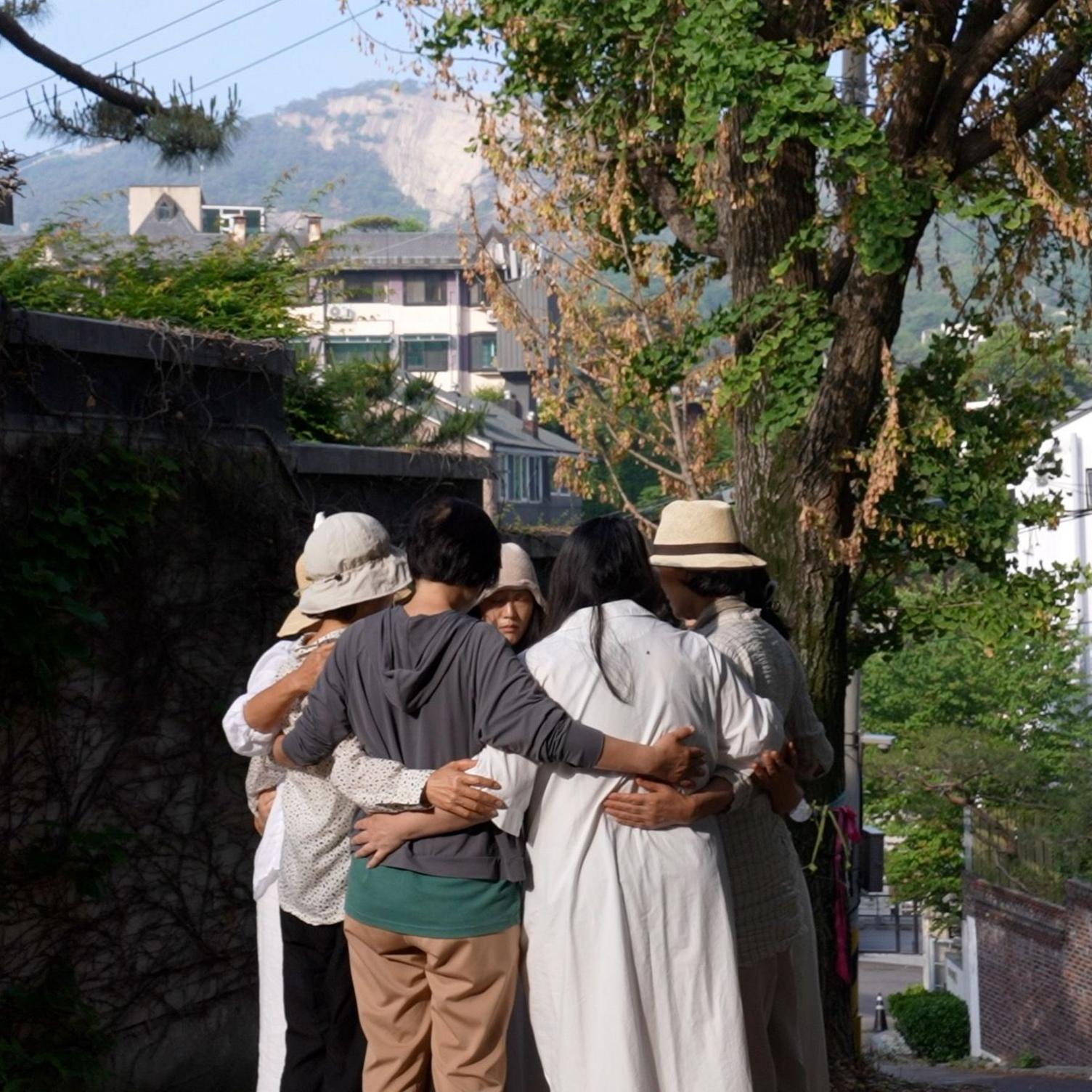 Several people standing outdoors beneath tall trees, arms around each other in a circle.