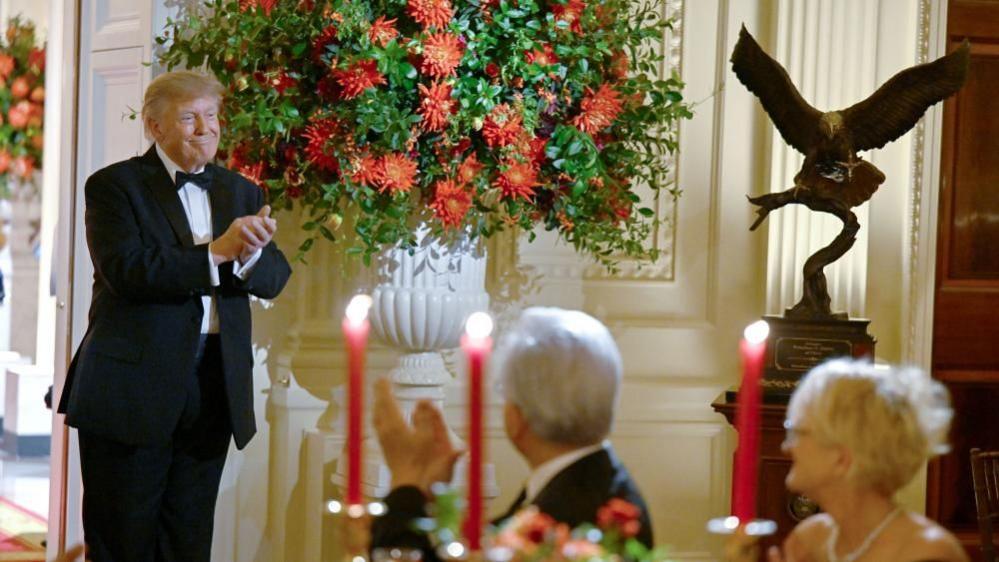 Trump, in a tuxedo, stands smiling next to a large white vase filled with red flowers. To the left is a large bronze statue of an eagle on a pedestal, with its wings outstretched.