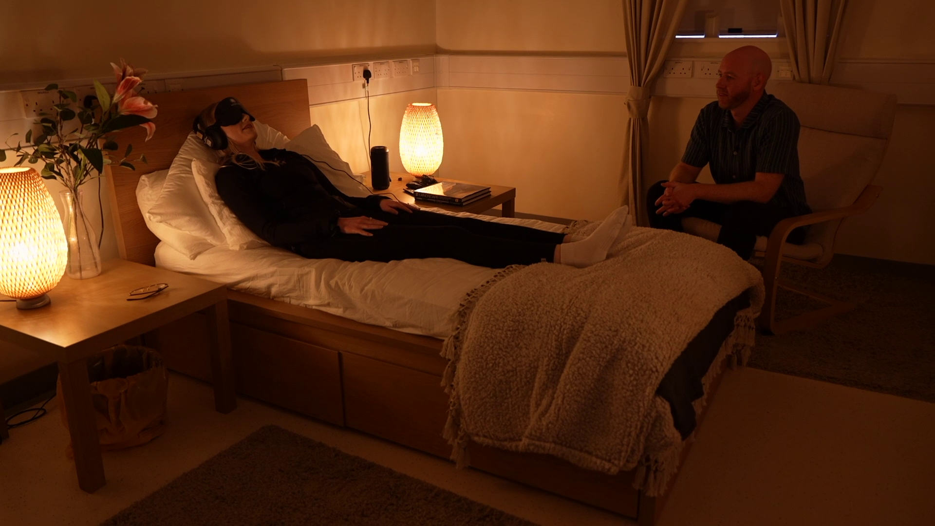 The dosing room at Care Pathway Enhancement (CaPE) Clinic. Showing a bedroom-like setting with a single bed with a wooden frame, there is a woman on the bed with a face mask and earphones on, the room is dimly lit by lamps on the wooden bedside table with a lounge chair at the side of the room with the facilitator Tom Sharp.