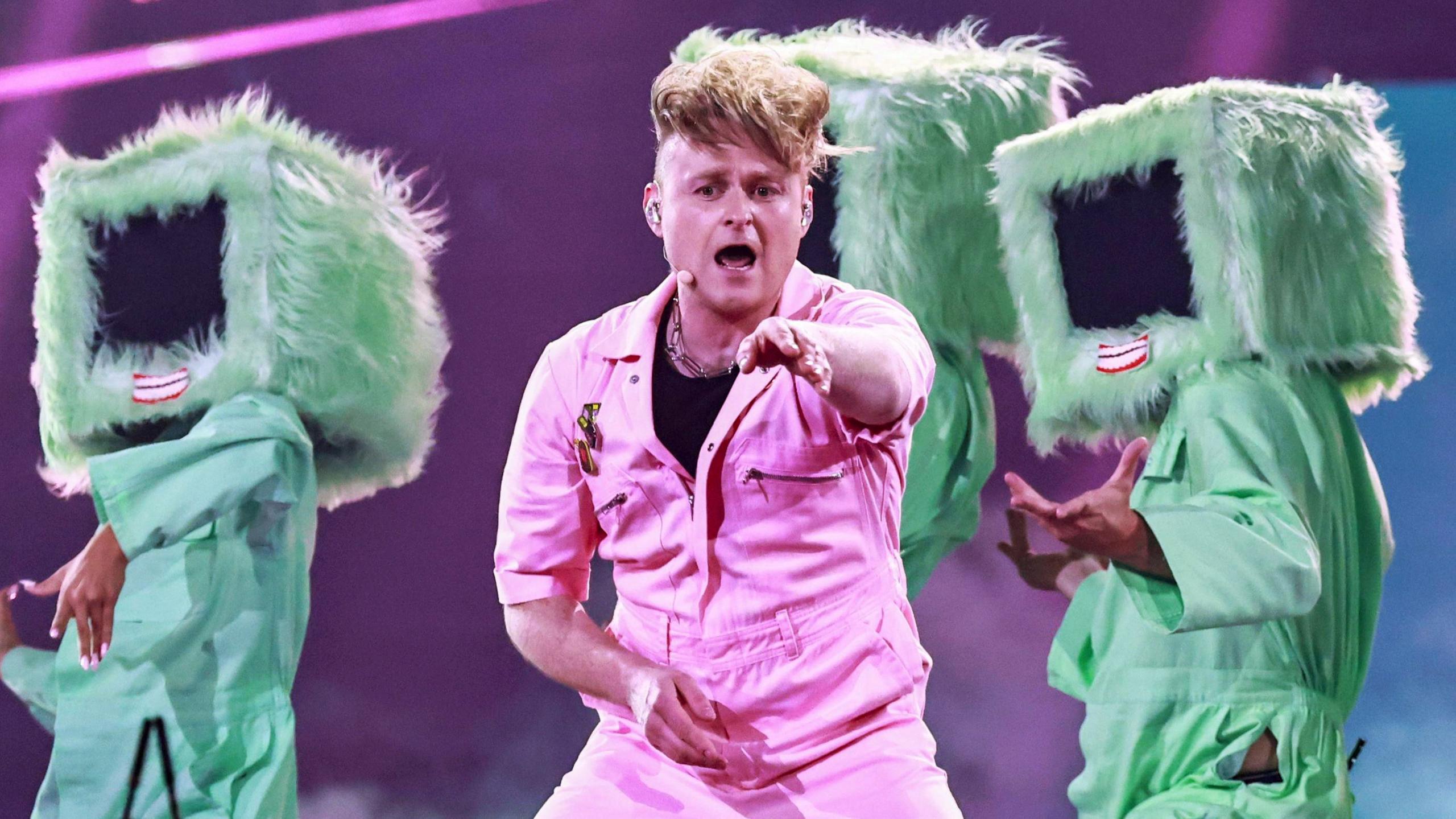Sam Battle aka Look Mum No Computer performing on stage during Eurovision dress rehearsals in a pink jumpsuit pointing towards the camera, with three dancers behind him wearing fur-lined computer monitors with red lips over their heads