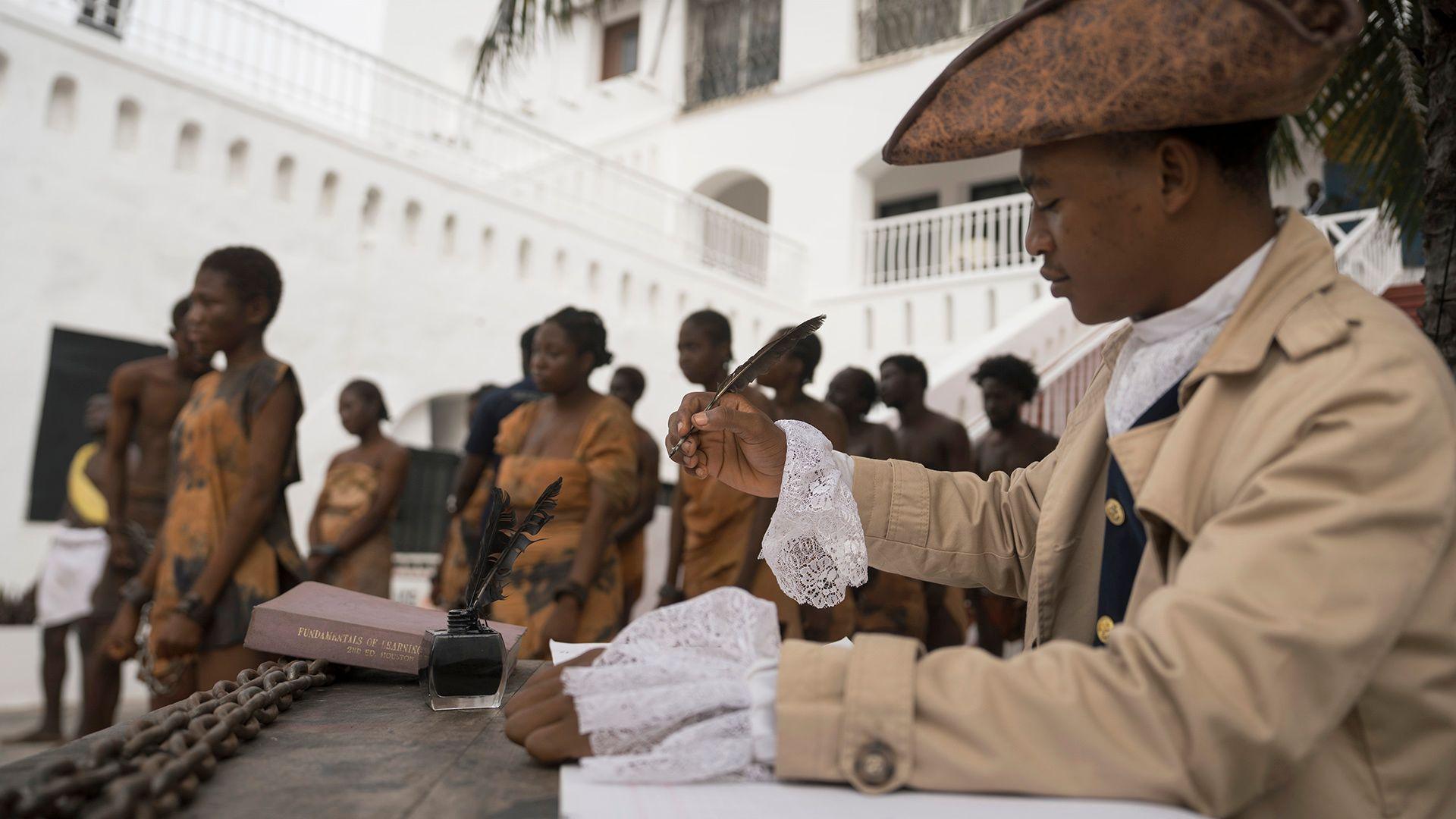 A person in the foreground wears pre- Victorian-era clothing and signs a document with a quil while wearing a hat, as people dressed in scantily clad slave robes walk through the street in chains in the background, during a re-enactment of the trans-Atlantic slave trade in the courtyard of Christiansborg Castle in Accra, Ghana on 19 June.