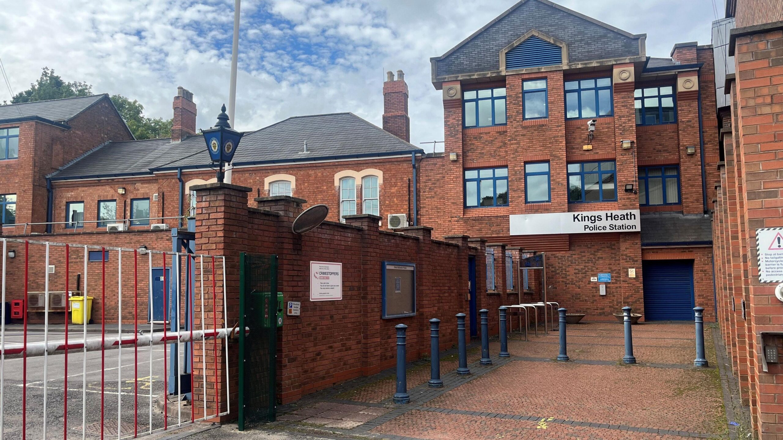 A red brick modern building with a courtyard in front, with blue bollards. A silver sign reads Kings Heath police station