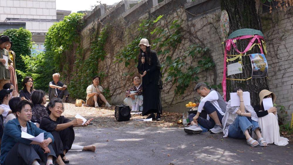 A group of about a dozen people sit and stand along a vine-covered brick wall. In the centre, a person dressed in black speaks into a microphone. To the right, a large tree is decorated with colorful ribbons, photographs, and papers.