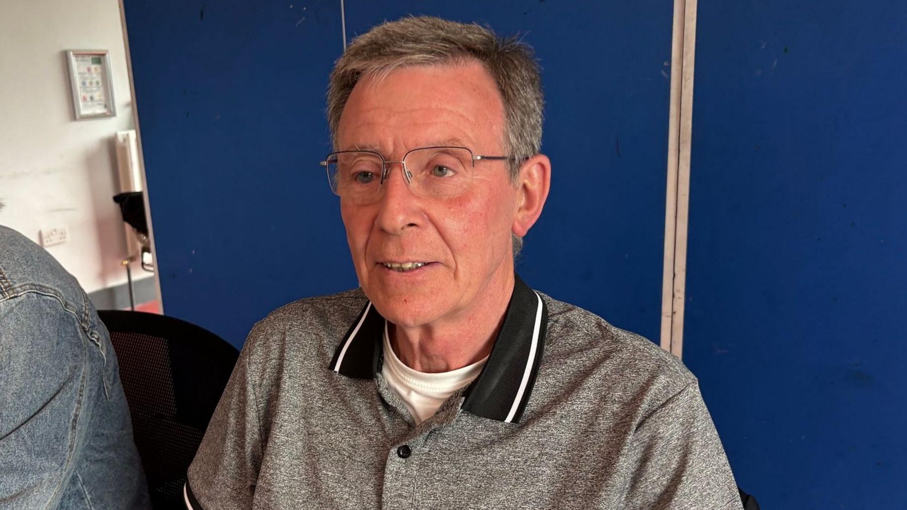Breandán Mac Cionnaith has short grey/brown hair parted to the side and is wearing wire-rimmed glasses