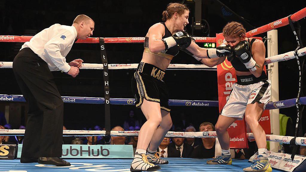 The referee looks on as Katie Taylor punches her opponent onto the ropes