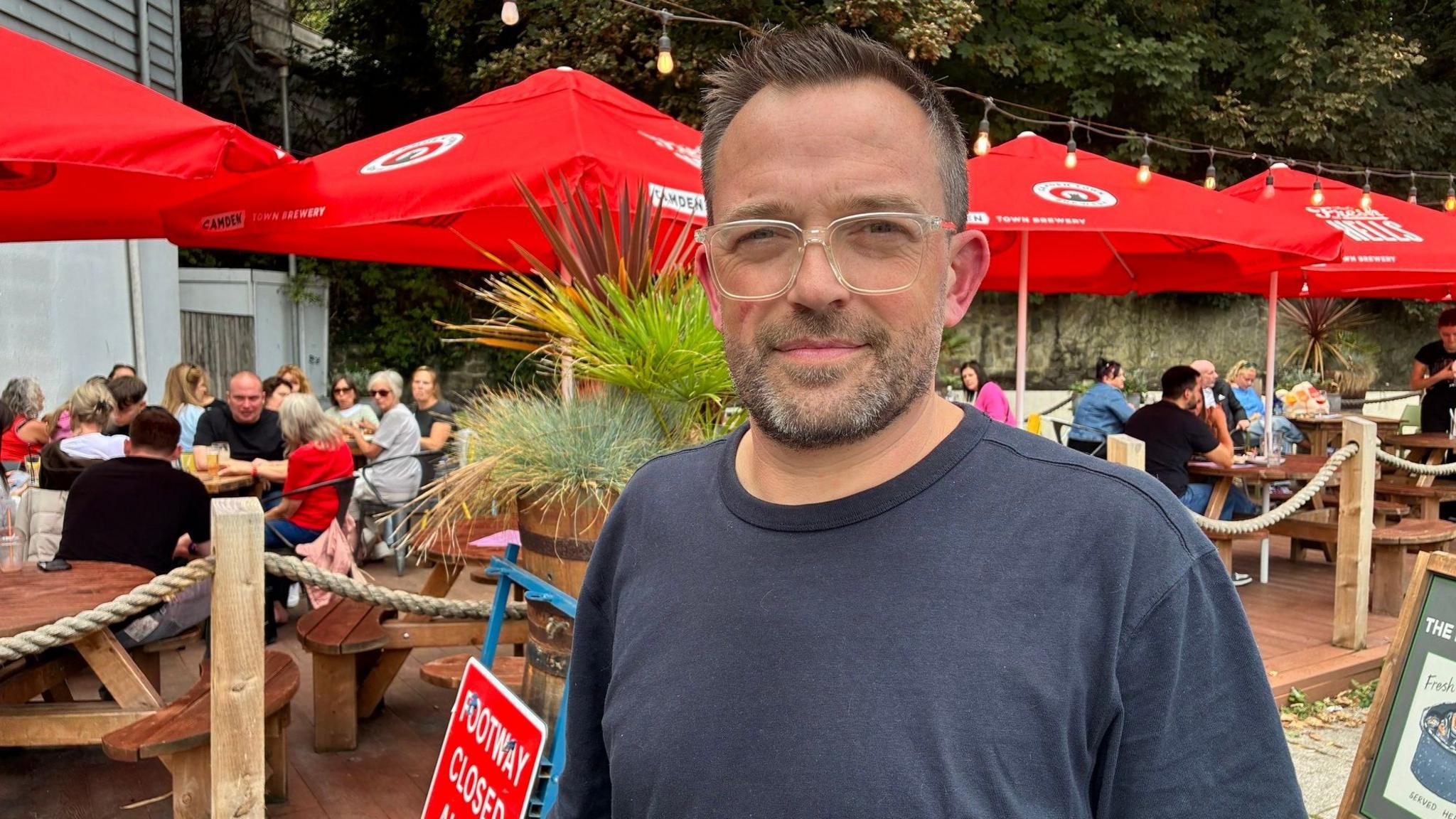 A man wearing glasses and a dark navy shirt. He is standing in front of an alfresco dining area.