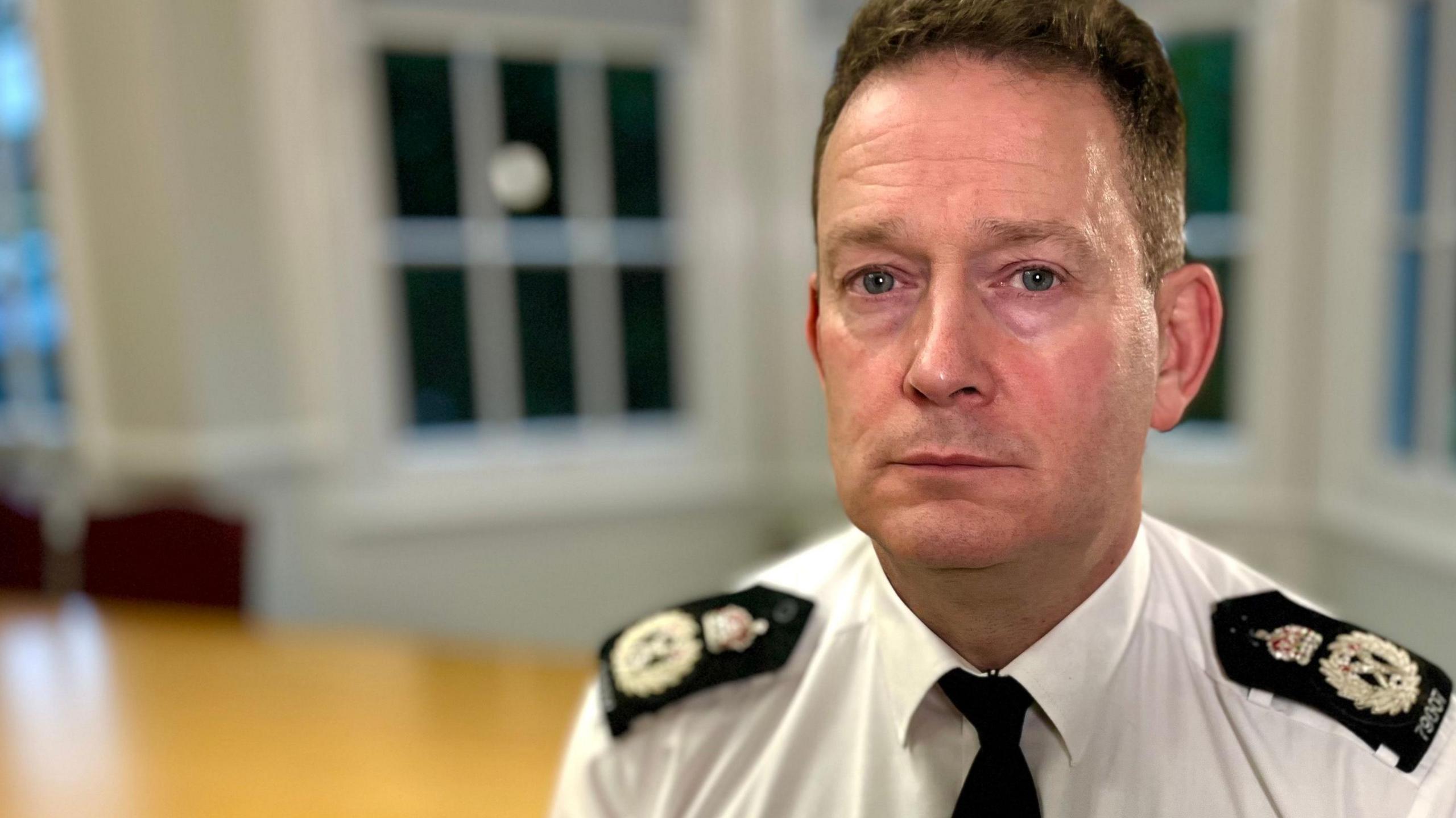 Ben-Julian Harrington sitting in a room with a desk and bay windows behind him. He wears a white shirt and black tie, and black epaulettes on his shoulders.