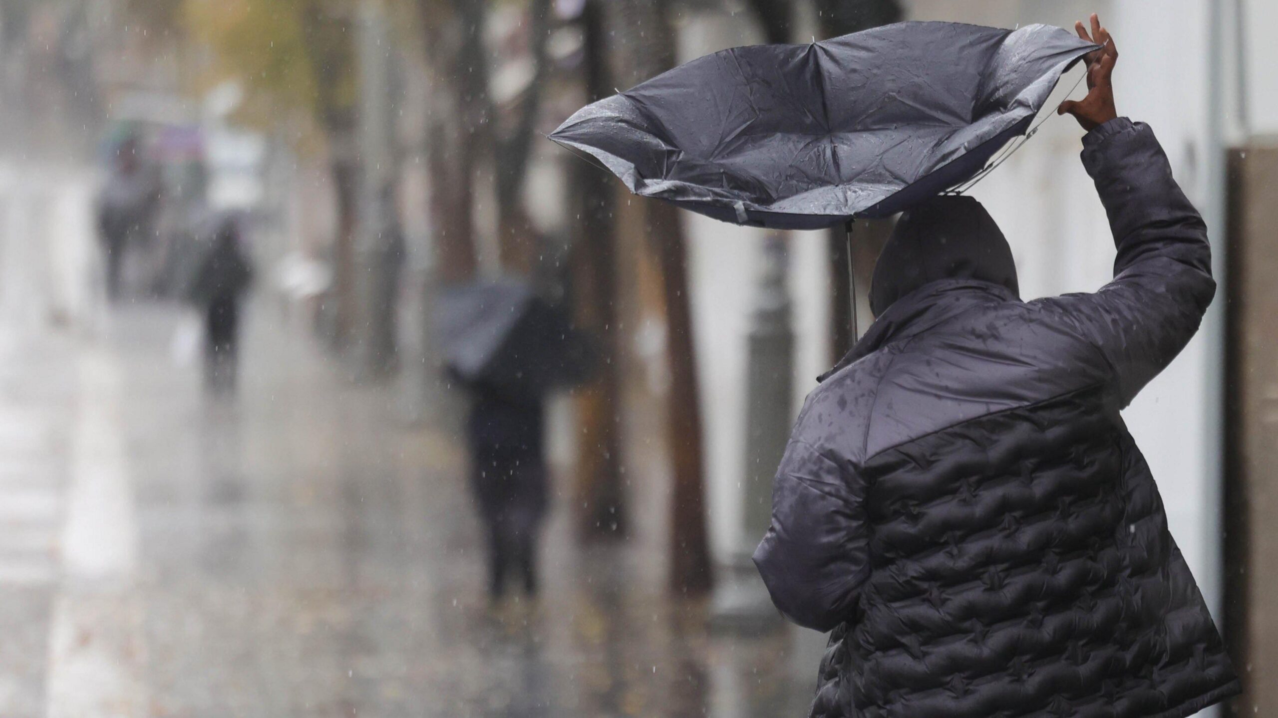 A man walks along the street in heavy rain. He holds a black umbrella which has turned inside out in the wind. Other people in the distance are also carrying umbrellas.