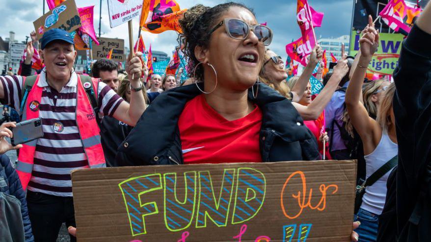 A large group of teachers and education staff on strike are gathered in a street raising their hands and holding flags and placards. In the foreground, a woman with dark hair tied back, hoop earrings and sunglasses, holds a sign that says "fund our schools". She wears a red t-shirt under a black coat. Behind her, a man wearing a blue hat, striped top and a high-vis pink vest holds a flag.