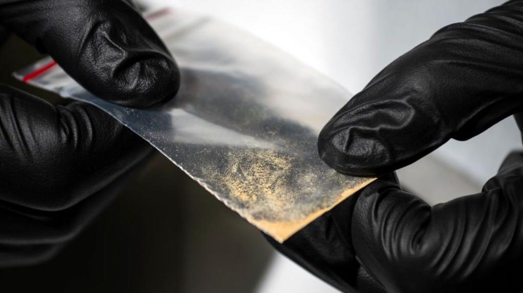 Close-up of gloved hands holding a small transparent plastic packet containing a light-coloured powdery substance. The packet is being examined against an out-of-focus laboratory background.