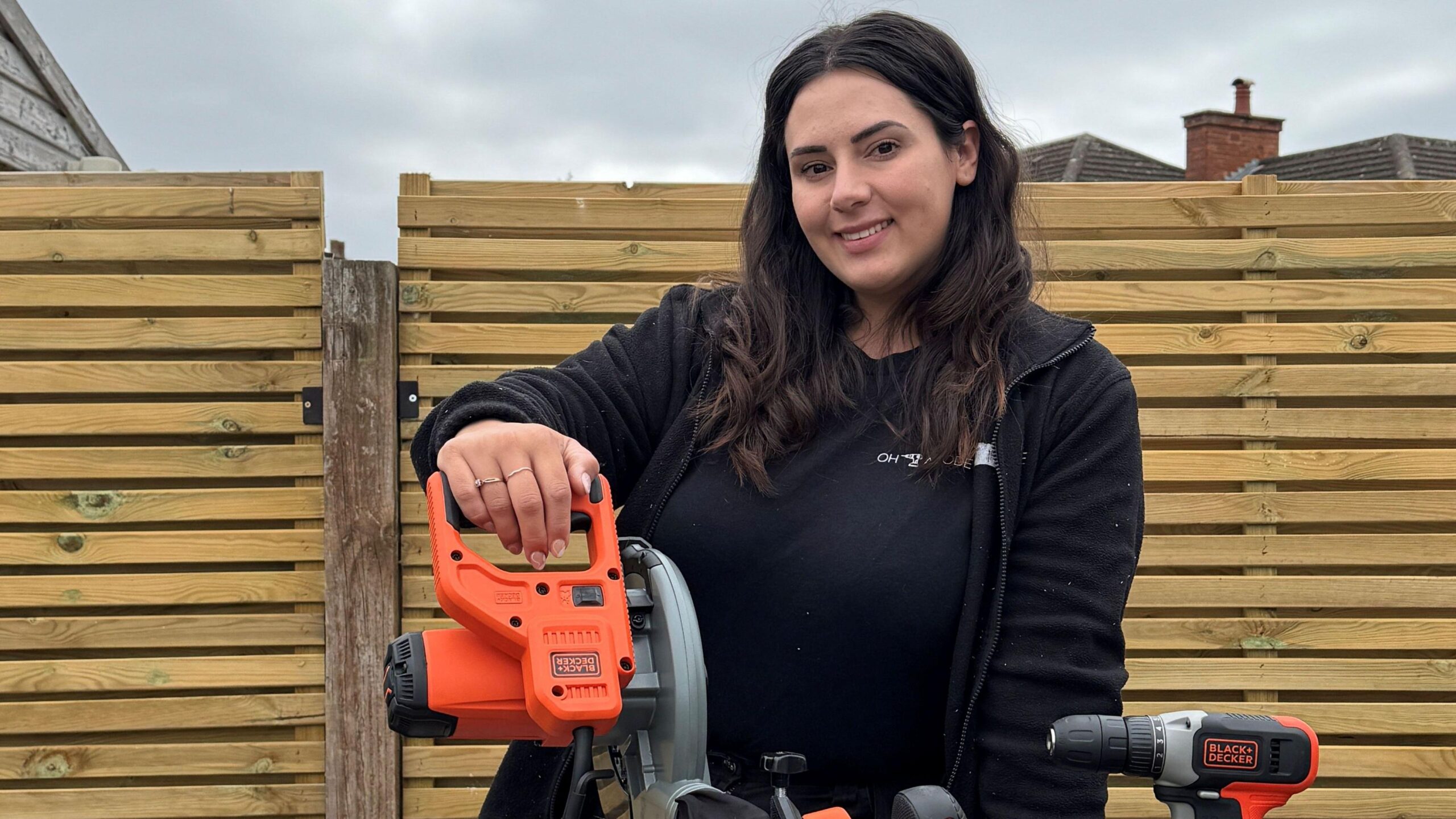 DIY influencer Jasmine Gurney stands outside in front of a fence, holding an orange and black power tool.