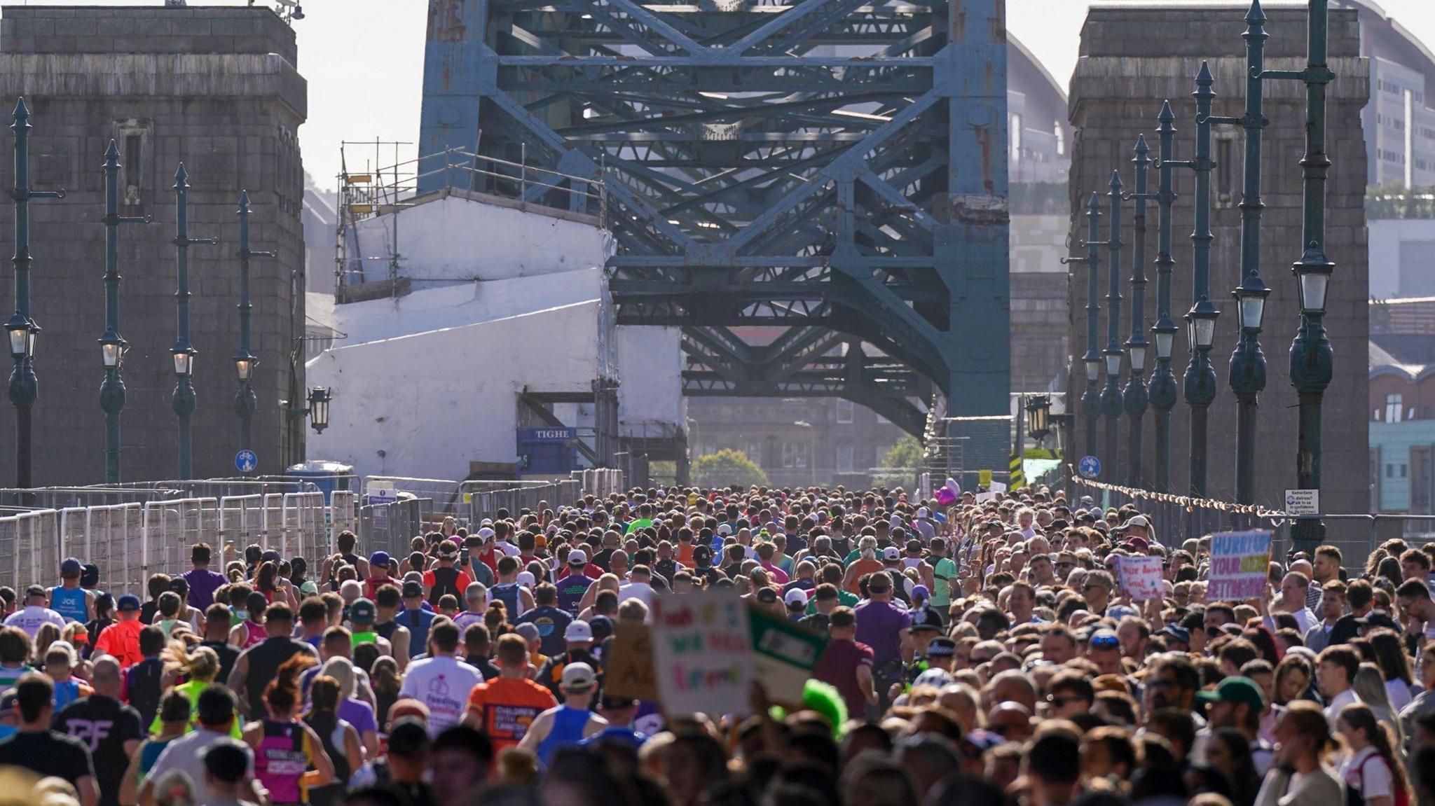 The backs of hundreds of runners running across the Tyne Bridge which is partially covered by scaffolding on the left. Spectators stand on the right, many holding up colourful signs and looking to the runners on the left.