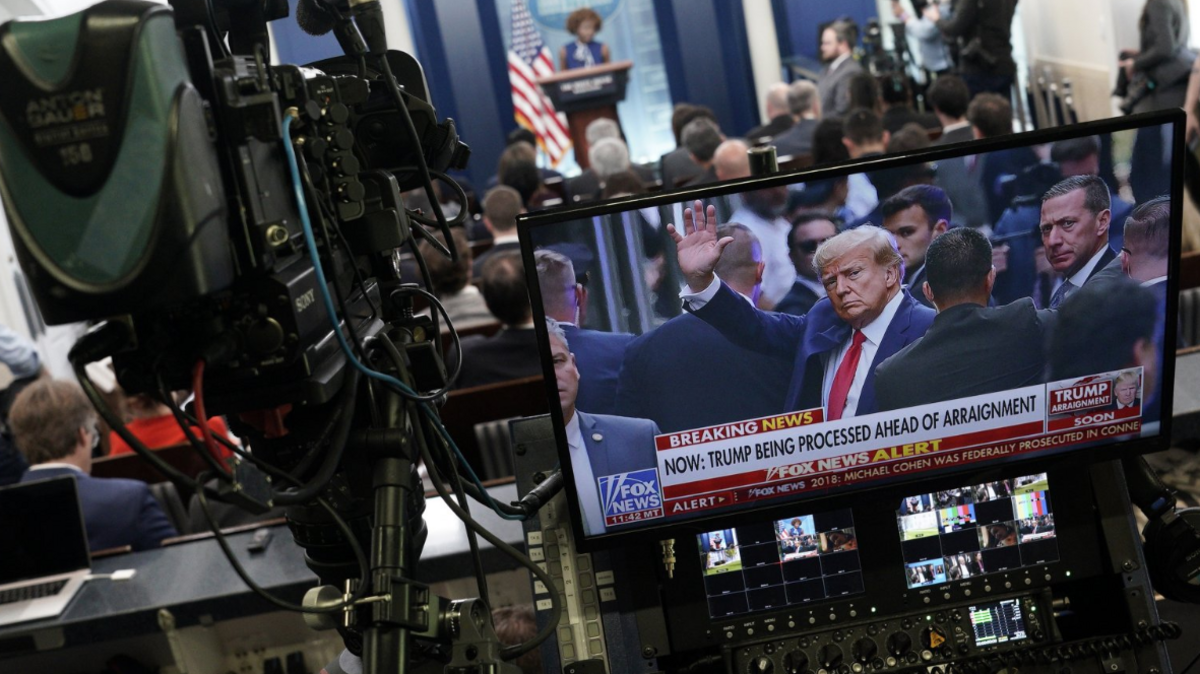 A press pool camera at a White House news briefing next to a screen showing Donald Trump, 4 April 2023.
