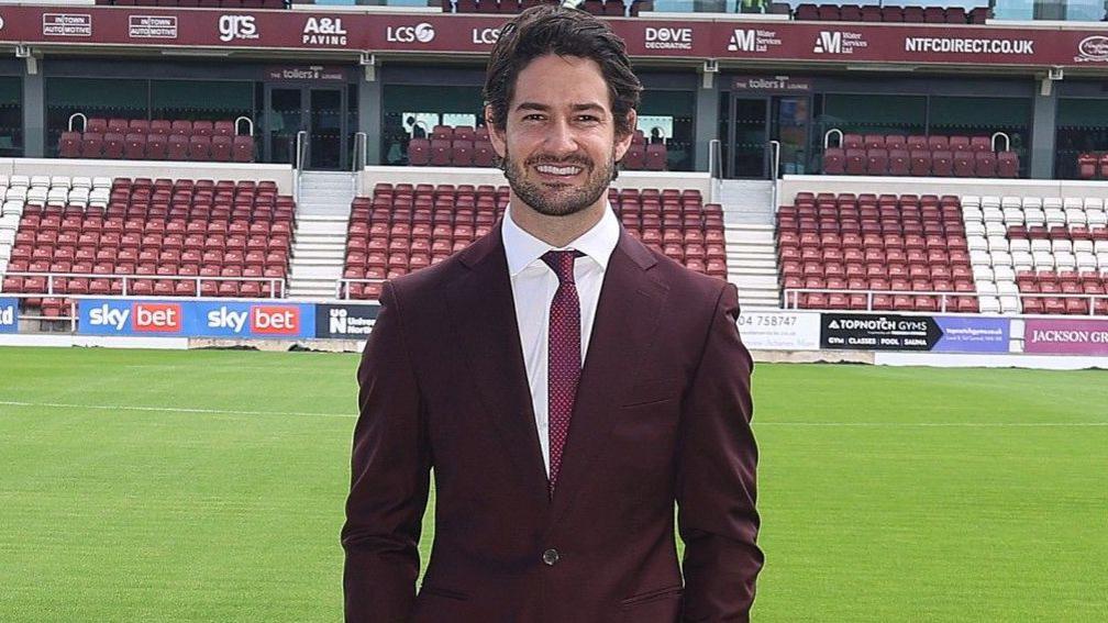 Alexandre Pato standing by side of Sixfields pitch, wearing maroon suit, white shirt, red tie and cream shoes, smiling with hands in pockets.