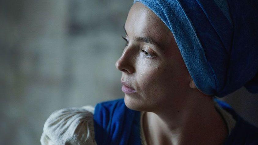 Side view of Jodie Comer. She is wearing a blue head wrap, a beige dress with a blue apron.