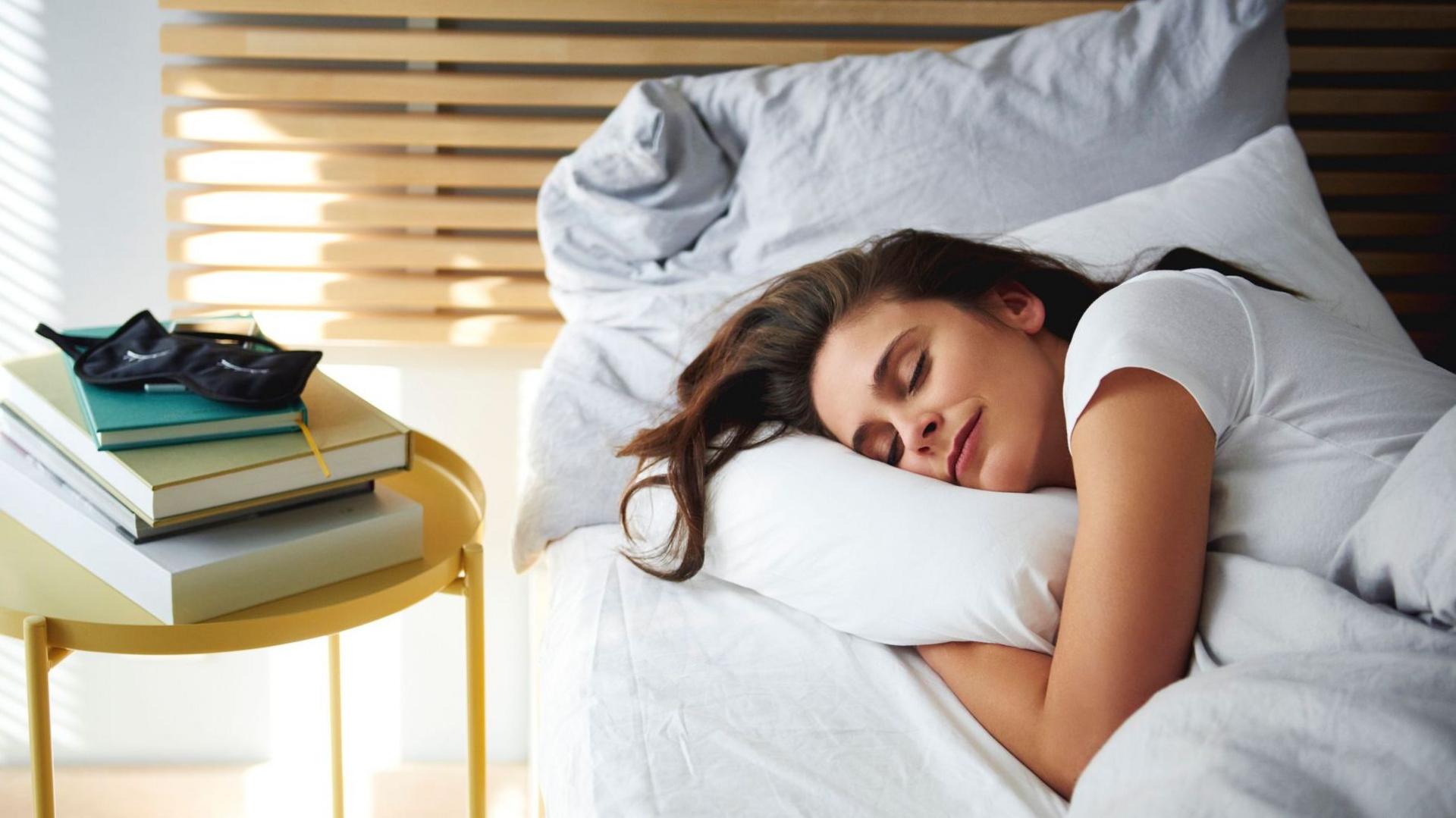 A young woman sleeping in a bed with white sheets and a gold night stand next to her with books and a sleep mask on