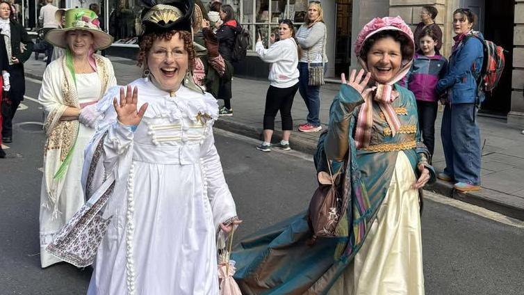 Bonny Wise and her friends are dressed in Regency-era dresses and are waving at the camera. They are on a road in Bath that is filled with people. They are all smiling.