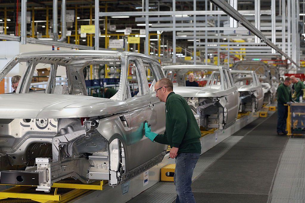 A factory line of shells of cars- a man wearing a green jumper, safety goggles and a green glove is standing sideways working on one of the cars. Colleagues can be seen in the background also wearing green jumpers.