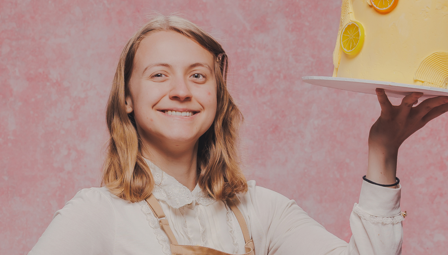 Bake-Off promo studio shot of Danni standing in a pink studio holding up a yellow iced cake on a board with one hand. They have shoulder length dark blonde hair and are wearing a long sleeved blouse with a lace collar and a beige apron.
