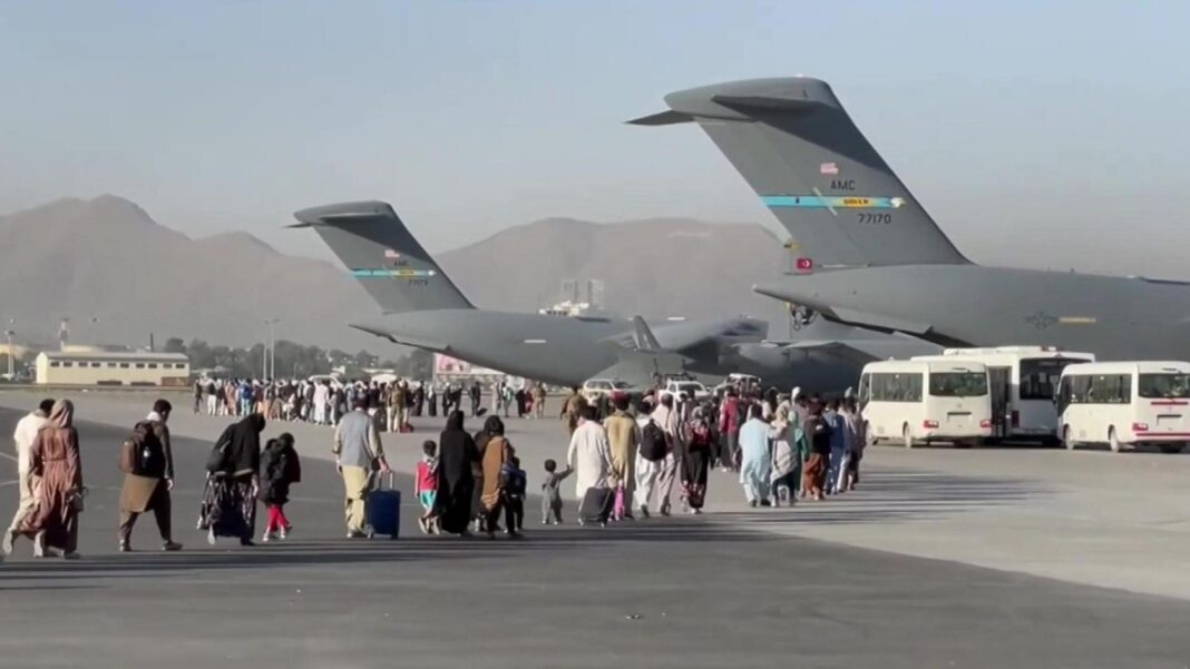 People carrying luggage walk across an airport tarmac towards large US military transport aircraft during an evacuation