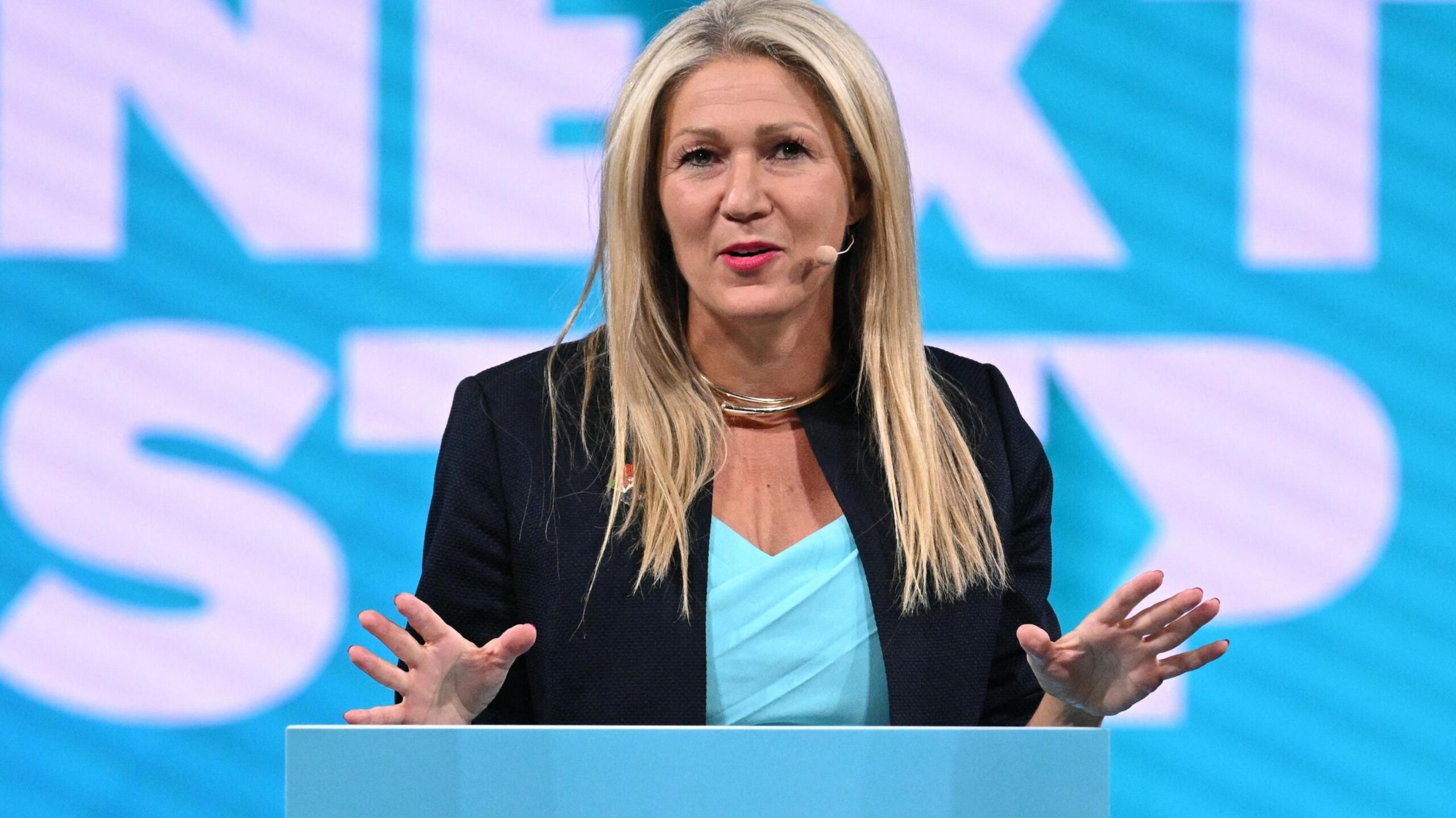 Laura Anne Jones stood at a podium in front of the words "Next Step" in Reform fontsetting and teal colours.
