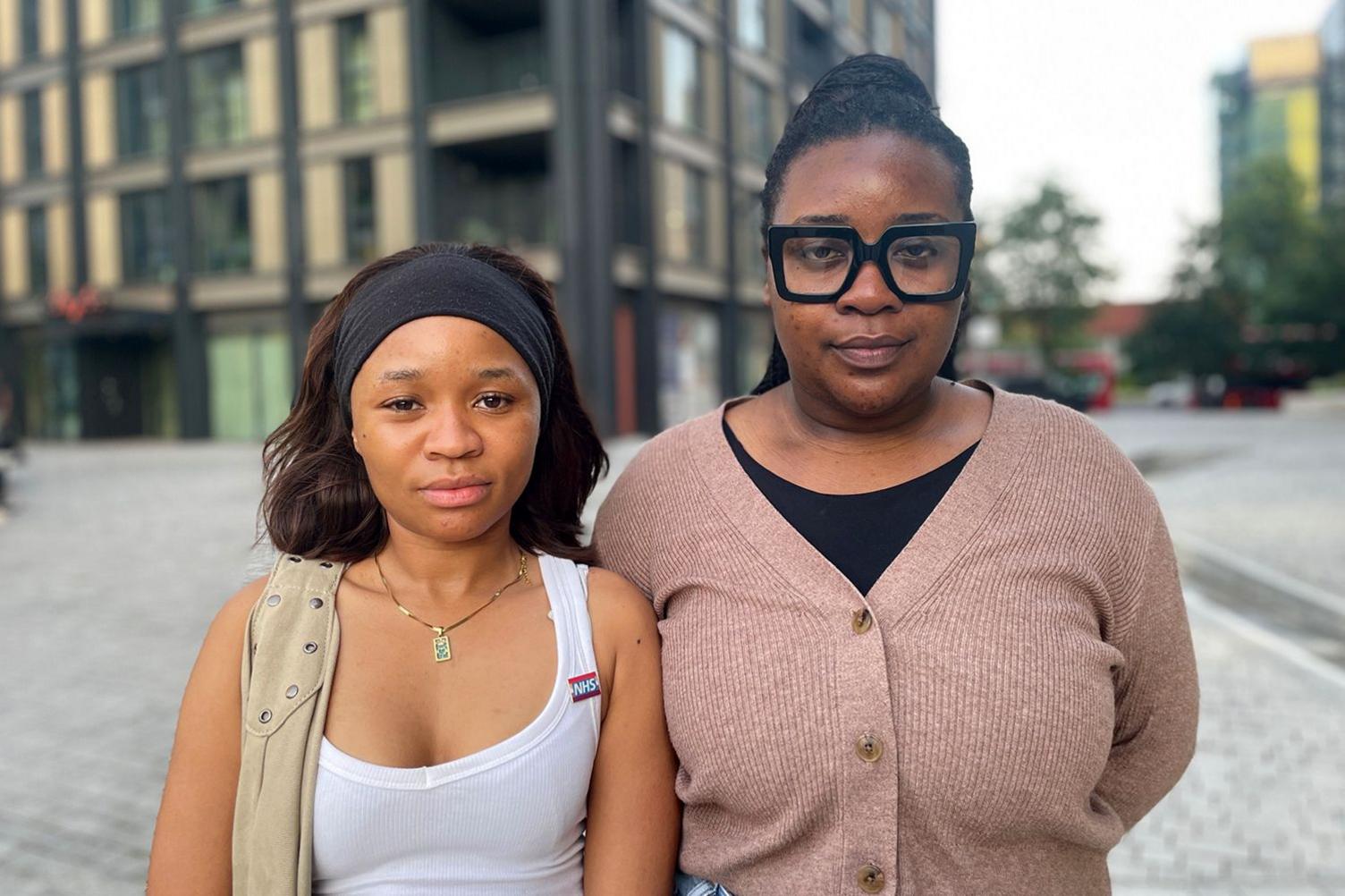 Two people stand outdoors on a paved area with modern buildings behind them. Sellah wears a white tank top with an NHS badge, a beige vest, a black headband and a gold necklace. Mphango wears large black‑framed glasses and a light brown cardigan over a black top.