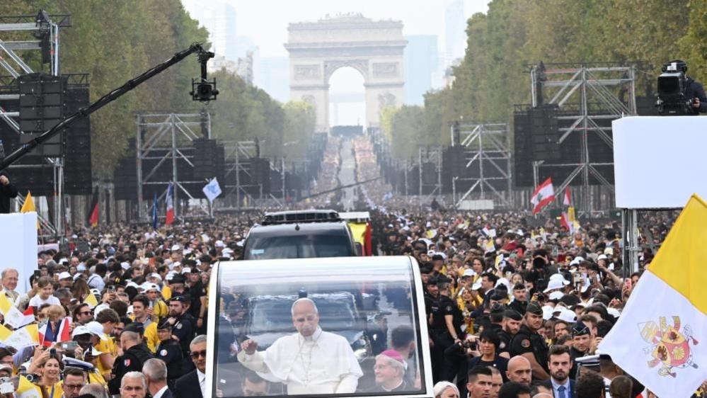 The pope can be seen in the centre of his popemobile. Champs-Élysées Avenue stretches out behind him and the Arc de Triomphe can be seen in the background. Crowds can be seen on either side of the road as well as screens which have been put up using scaffolding.