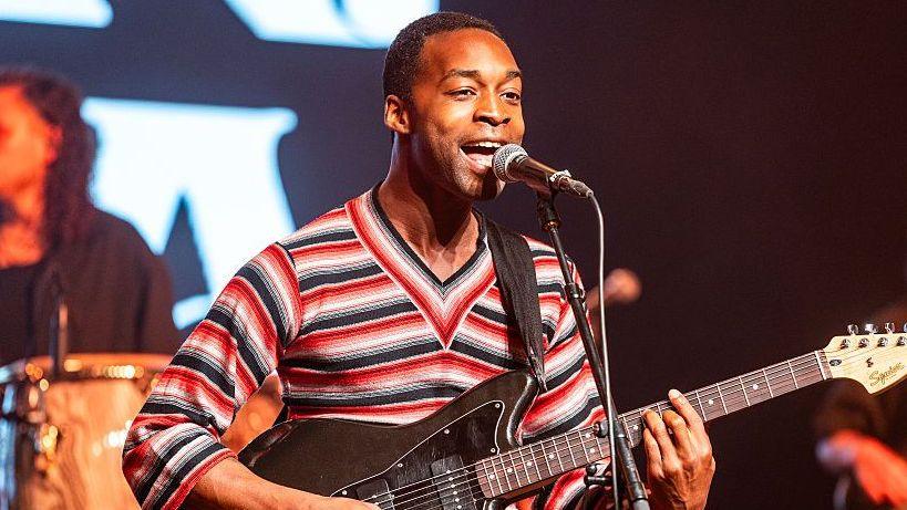 Jalen Ngonda performs on stage at a festival, wearing a red and black striped v-neck top. He plays his guitar which is strapped across his chest and looks out to the crowd. A drummer from his band is seen in the background.