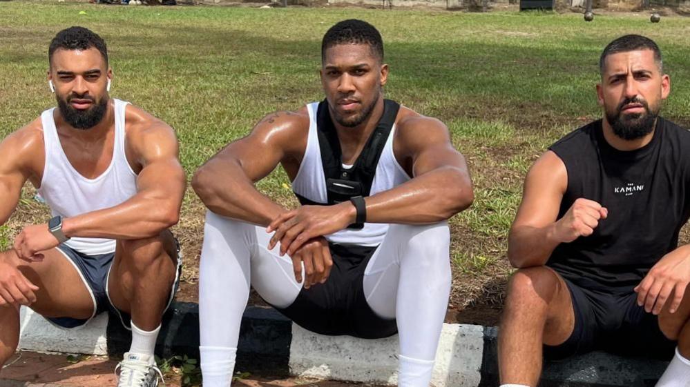Latif Ayodele sits with Anthony Joshua and Sina Ghami by grass outside, all in training gear