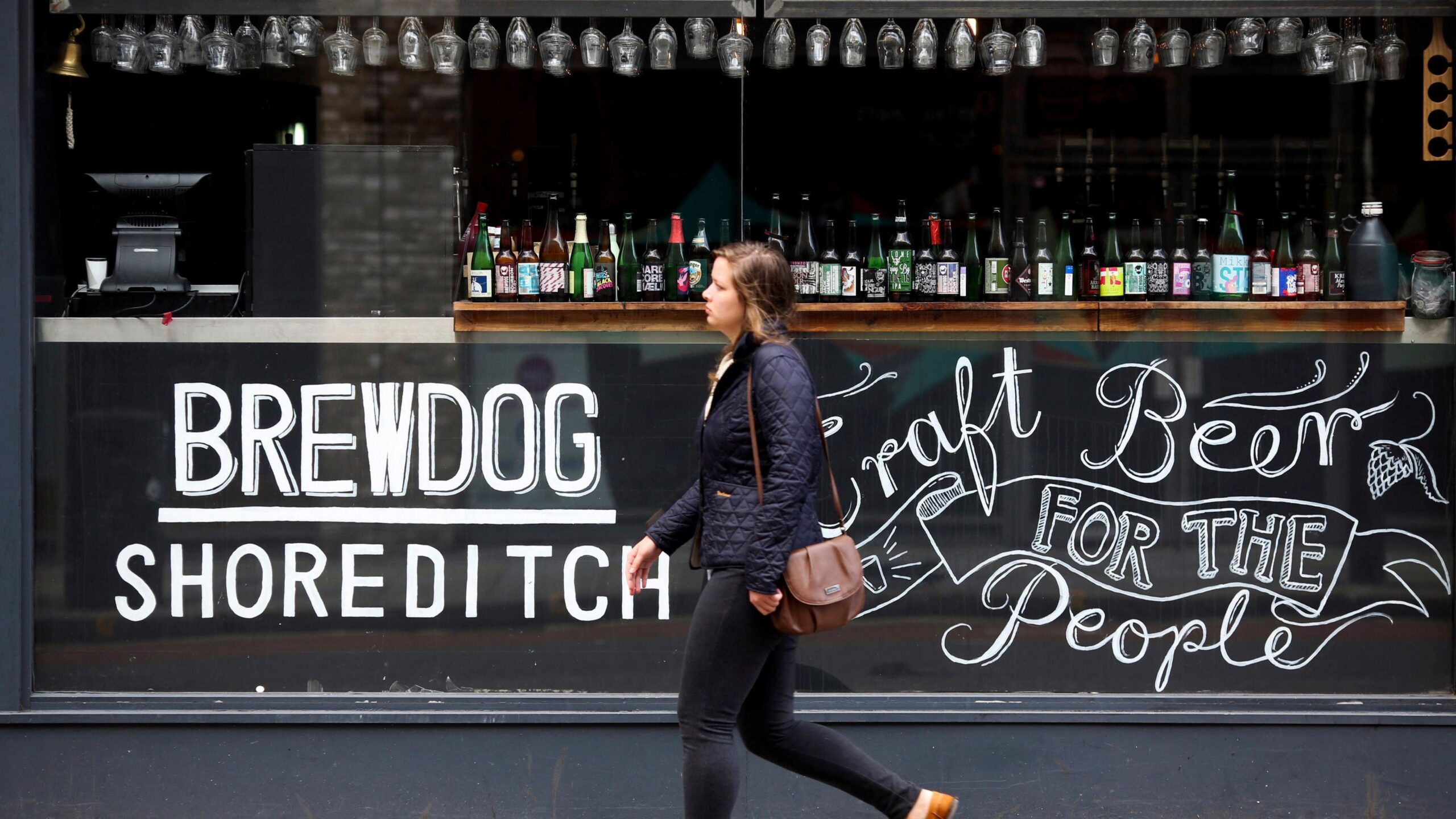 A woman walks past a Brewdog bar in London. On the glass is written Brewdog Shoreditch and Craft Beer for the People.