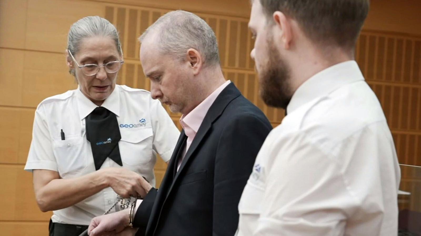 A uniformed female security officer adjusts handcuffs on Graeme Dott, who is wearing a dark suit jacket and light shirt in a wood-panelled courtroom.   Another uniformed security officer, with a beard, stands in the foreground with their back partly to the camera.