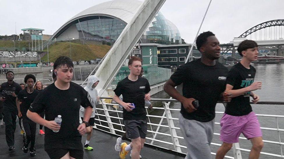 A group of young men and women are running over the Gateshead Millennium Bridge with it's white railings to one side and the white, curved metal support beginning to arch over them. Beyond is The Glasshouse music centre and further beyond is the Tyne Bridge, with the grey River Tyne beneath. The runners are mostly wearing black and grey but one one young man has bright fuchsia shorts.