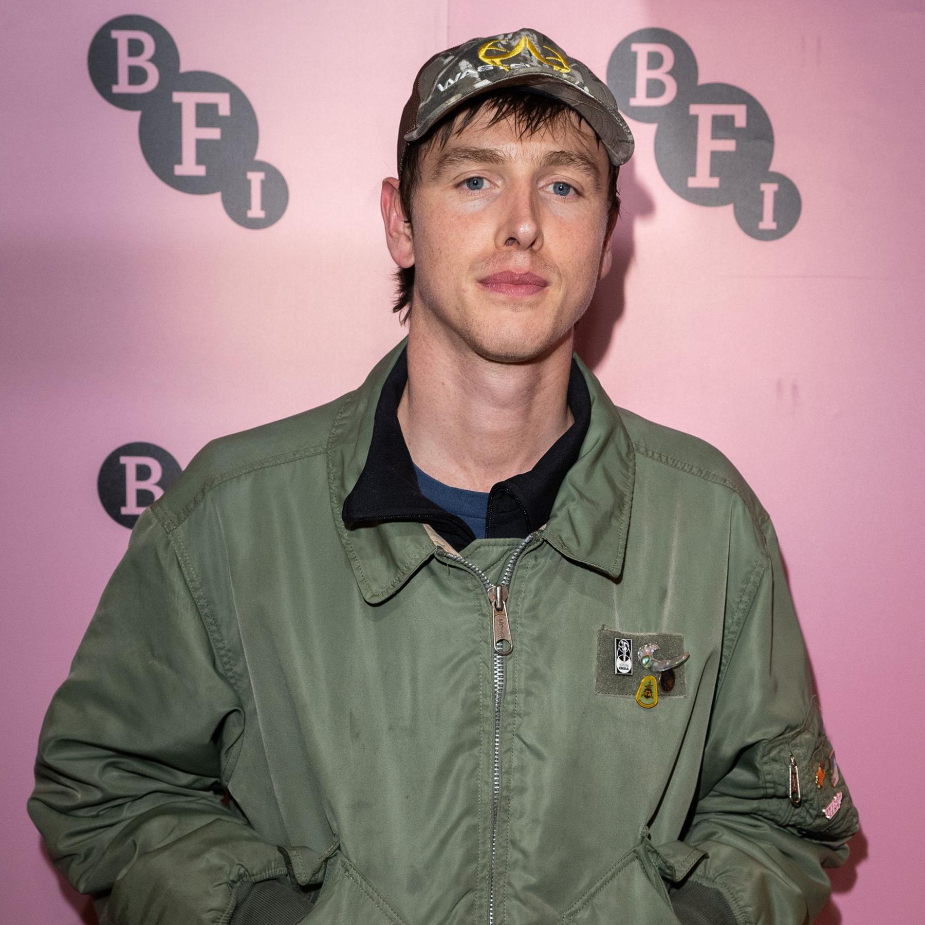 Harris Dickinson poses for a photo while wearing a cap and casual bomber jacket against a pink backdrop with the BFI logo visible, in London in 2025.