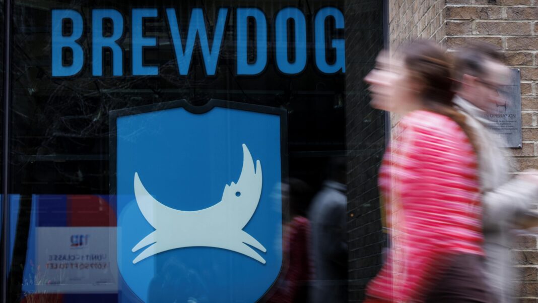 Brewdog’s unpaid workers to receive nothing after takeover deal People walk past a closed-down BrewDog bar in central London. The window of the pub has the word Brewdog written in large blue letters above its logo, featuring a white dog on a blue background.