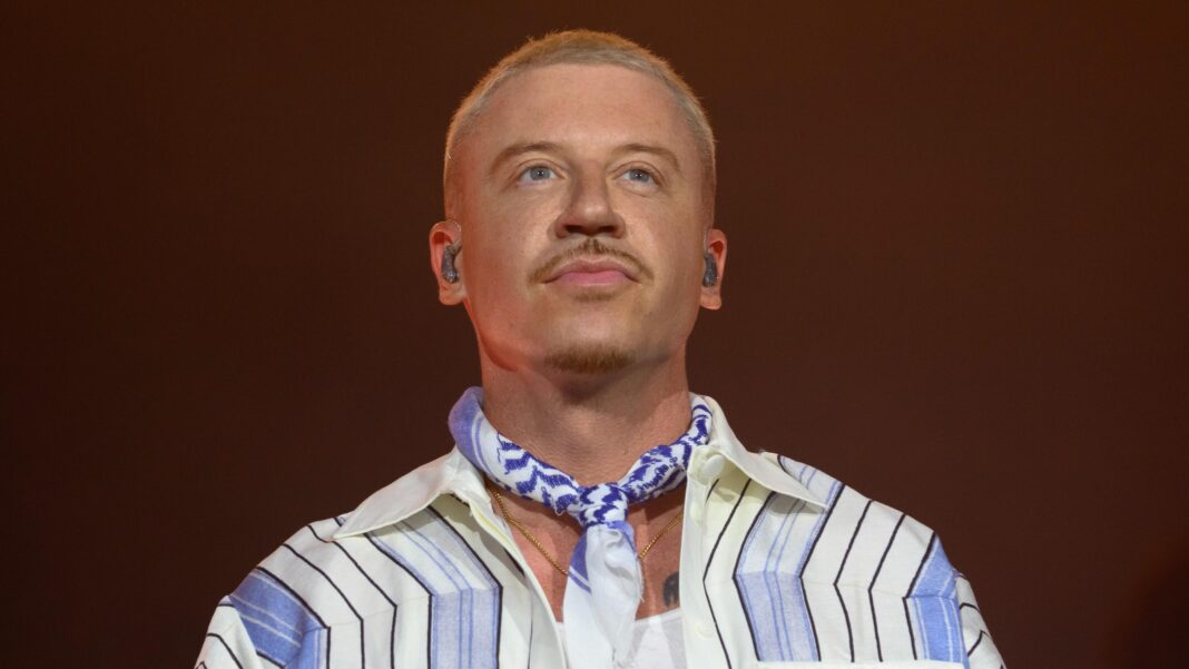 Close up of Macklemore, who has shaved blonde hair, and is wearing a blue striped shirt.