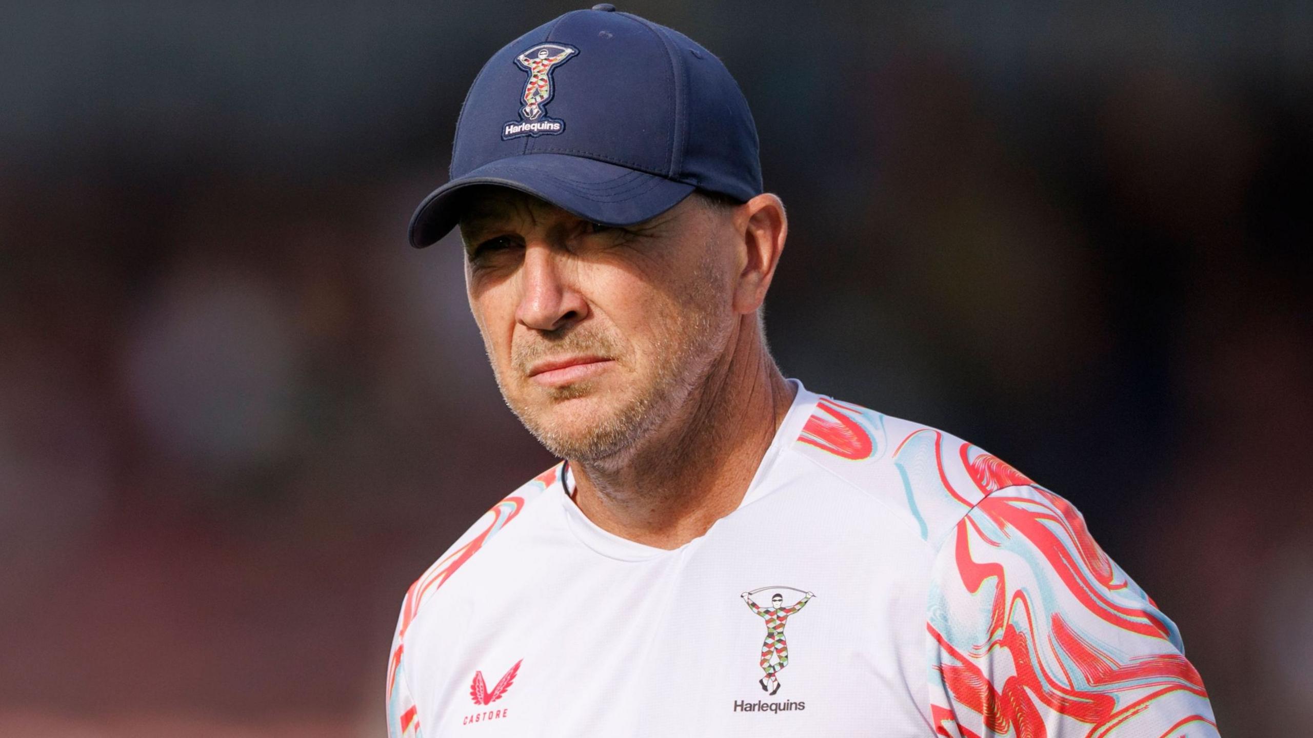Jason Gilmore wearing a navy blue Harlequins baseball cap and a white Harlequins training top with red and light blue patterns on the shoulders and sleeves