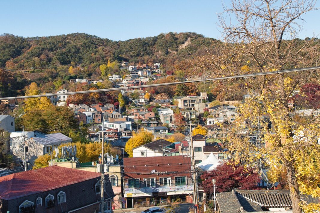 Panoramic view of a hillside neighbourhood in autumn, with clusters of houses and low-rise buildings nestled among colourful trees in shades of yellow, orange, red, and green. Forested mountains rise in the background under a clear blue sky, while a road and metal fence run across the foreground.